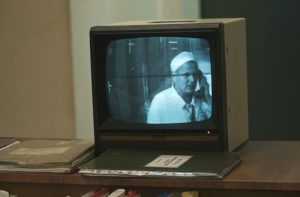 Kadr z filmu. Telewizor stojący na meblu, obok leżą teczki. Na ekanie widać czarno-biały kadr - mężczyzna w białym ubraniu i białym czepku rozmawia przez telefon stacjonarny.