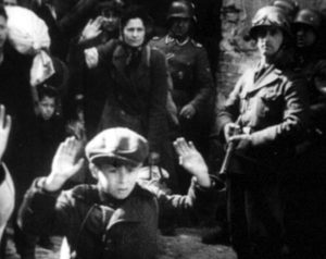 Kadr z czarno-białego filmu. Żołnierze trzymający broń. Na pierwszym planie chłopiec z uniesionymi w górę rękoma. W tle kobieta z uniesioną ręką.
