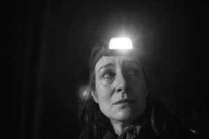 Kadr z filmu, czarno-biały. W ciemności widać twarz kobiety z latarką czołową na głowie.