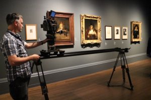 mężczyzna a kamerą na szynie, w tle obrazy Rembranta w złotych ramach