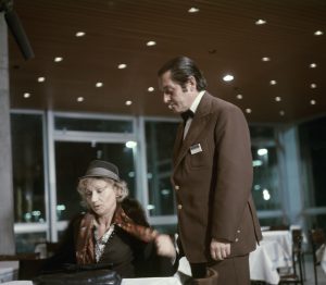 kobieta w kapeluszu, eleganckim płaszczu, siedzi przy stoliku. nad nią stoi mężczyzna w brązowym garniturze.